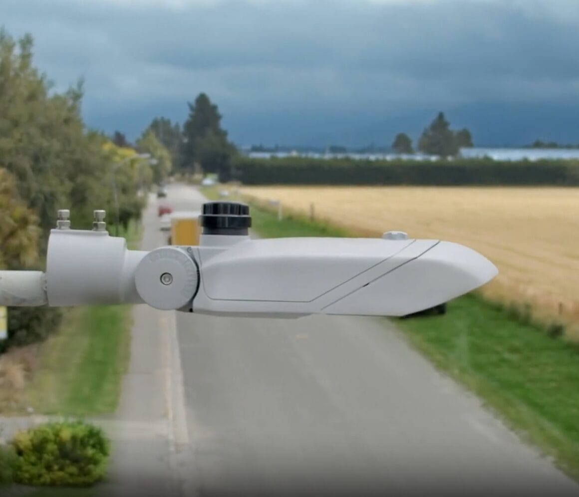 A close-up of a mounted weather sensor overlooking a rural road, with fields and trees on both sides under a cloudy sky.