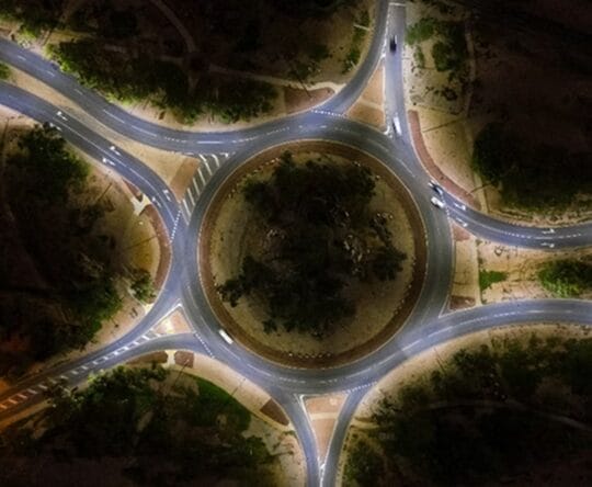 Aerial view of a roundabout at night, illuminated by streetlights, with several cars on the connecting roads.