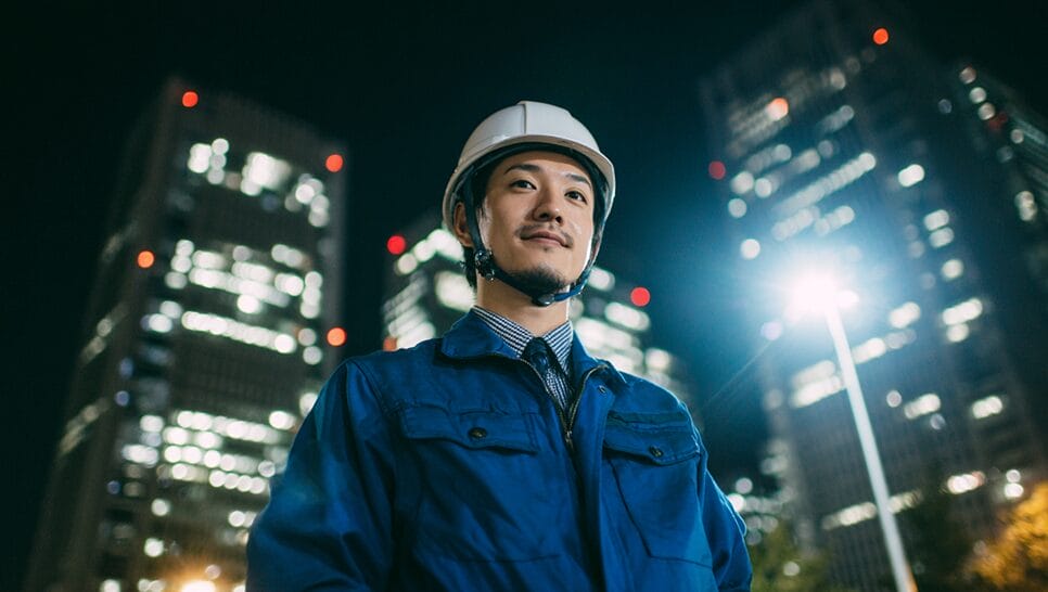 A man in a blue work jacket and white hard hat stands outdoors at night in front of illuminated office buildings.