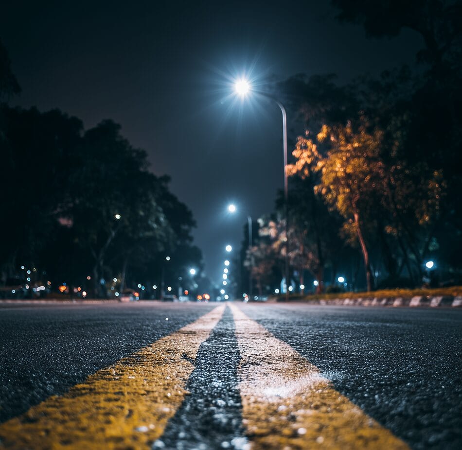 A dimly lit street at night, viewed from ground level with yellow double lines in the center, lined with trees and illuminated by streetlights.