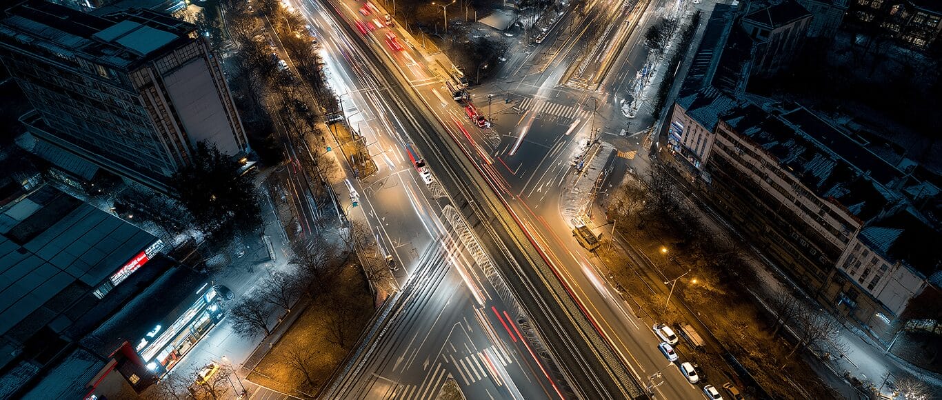 Aerial view of a busy city intersection at night with car light trails, illuminated streets, and surrounding buildings.