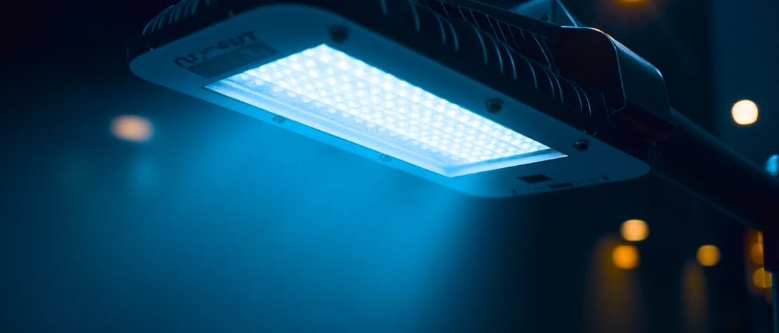 A close-up view of a modern LED streetlight illuminated with a blue glow, mounted on a pole against a blurred dark background.