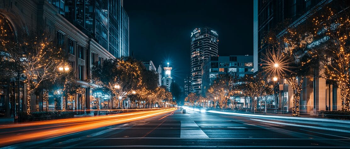 A city street at night with illuminated trees, tall buildings, and light trails from moving vehicles, creating a vibrant urban scene.