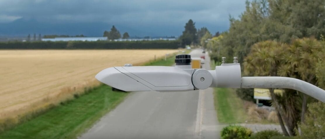 A modern LED streetlight is mounted on a curved pole overlooking a rural road, with fields and trees in the background under a cloudy sky.