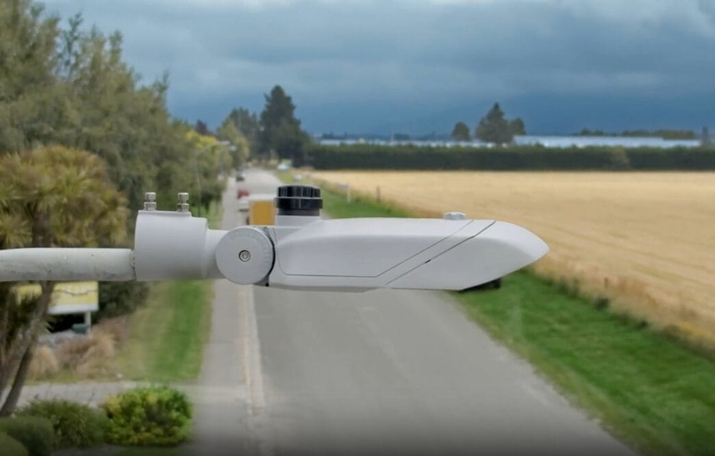 A close-up of a white weather sensor mounted over a rural roadway, with fields and trees on both sides under a cloudy sky.