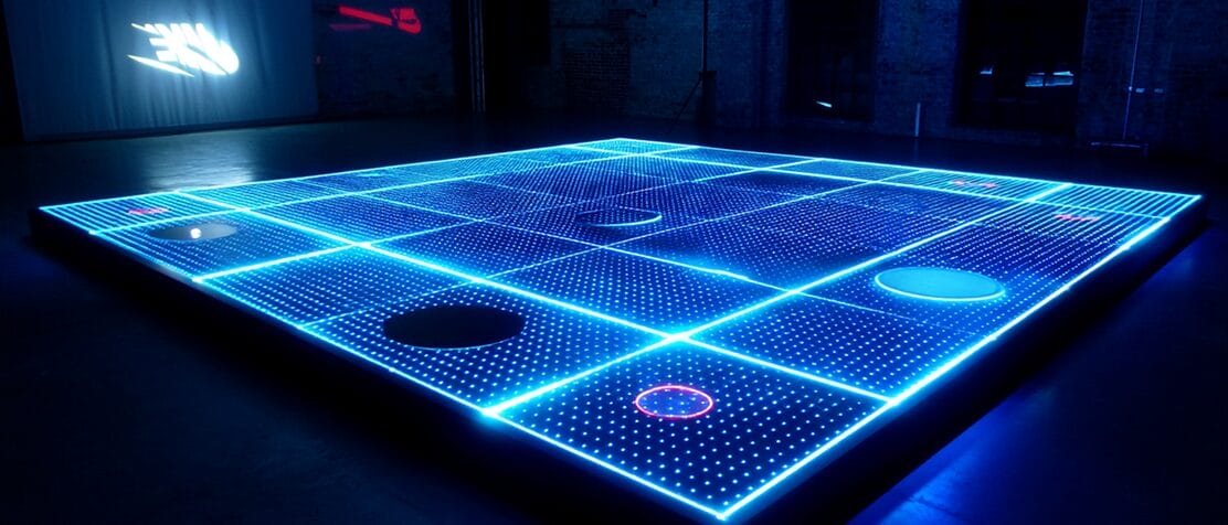 A large, illuminated grid floor with glowing blue lines and circles of varying colors on some squares, set in a dark indoor space.