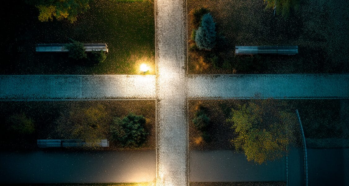 Aerial view of a cobblestone path intersection in a park at night, lit by streetlights, with benches and trees visible in each quadrant.