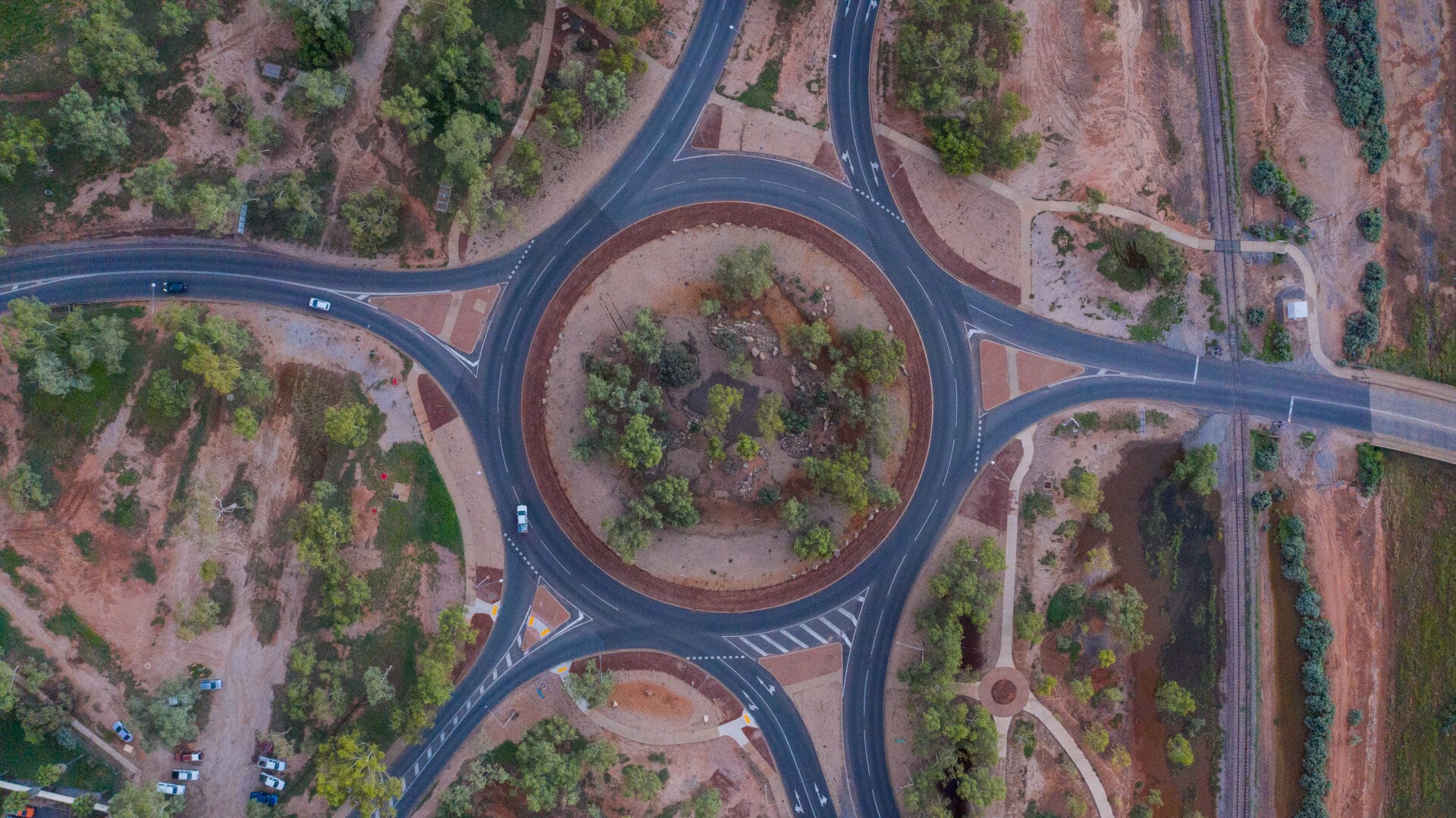 Aerial view of a circular traffic roundabout with multiple roads branching off, surrounded by greenery and dry terrain.
