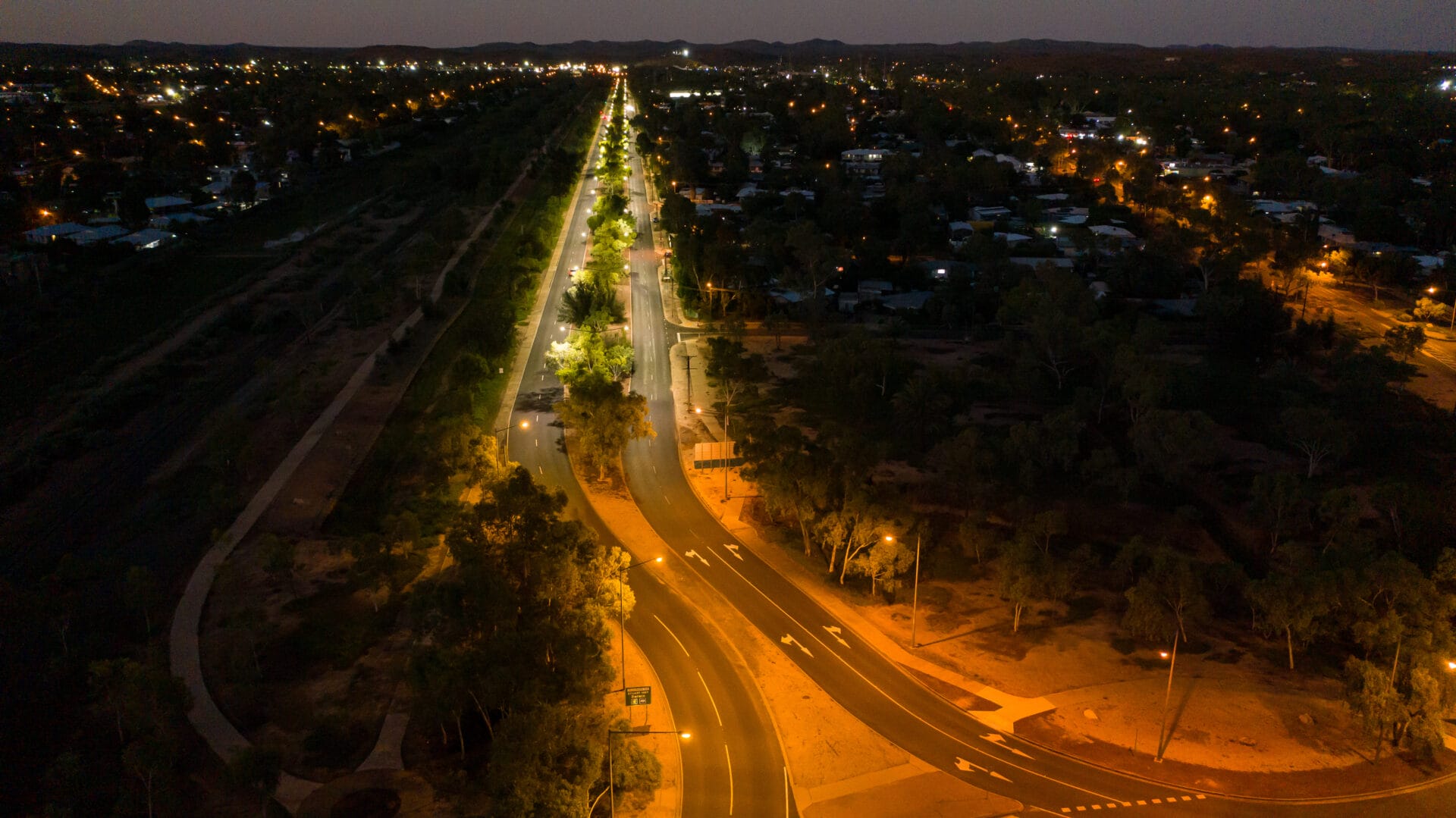 Aerial view of a straight, illuminated road stretching into the distance at night, with surrounding city lights and dark, tree-covered areas.