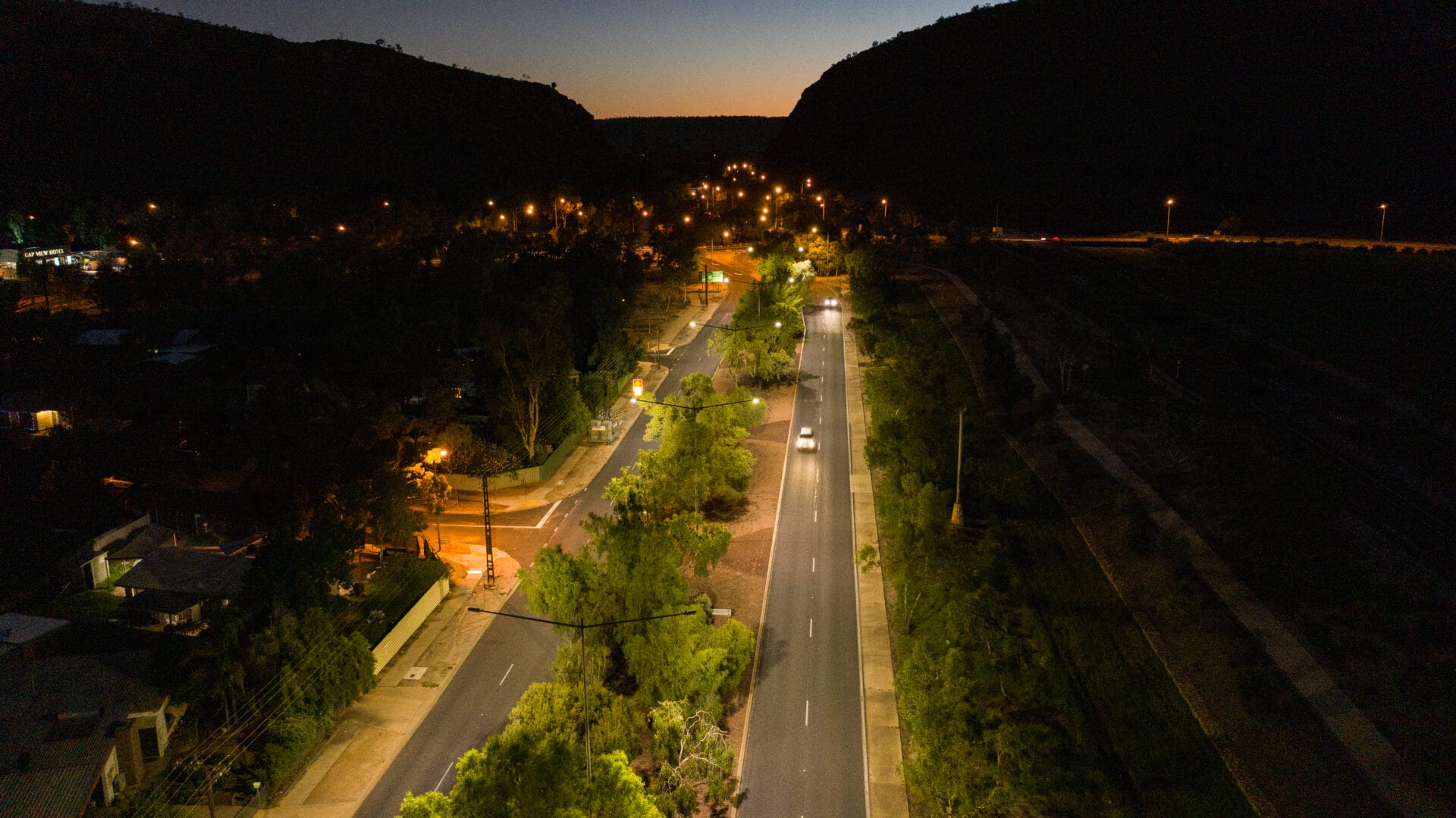 Aerial view of a dimly lit road at dusk, with streetlights illuminating the street and surrounding trees, flanked by hills in the background.