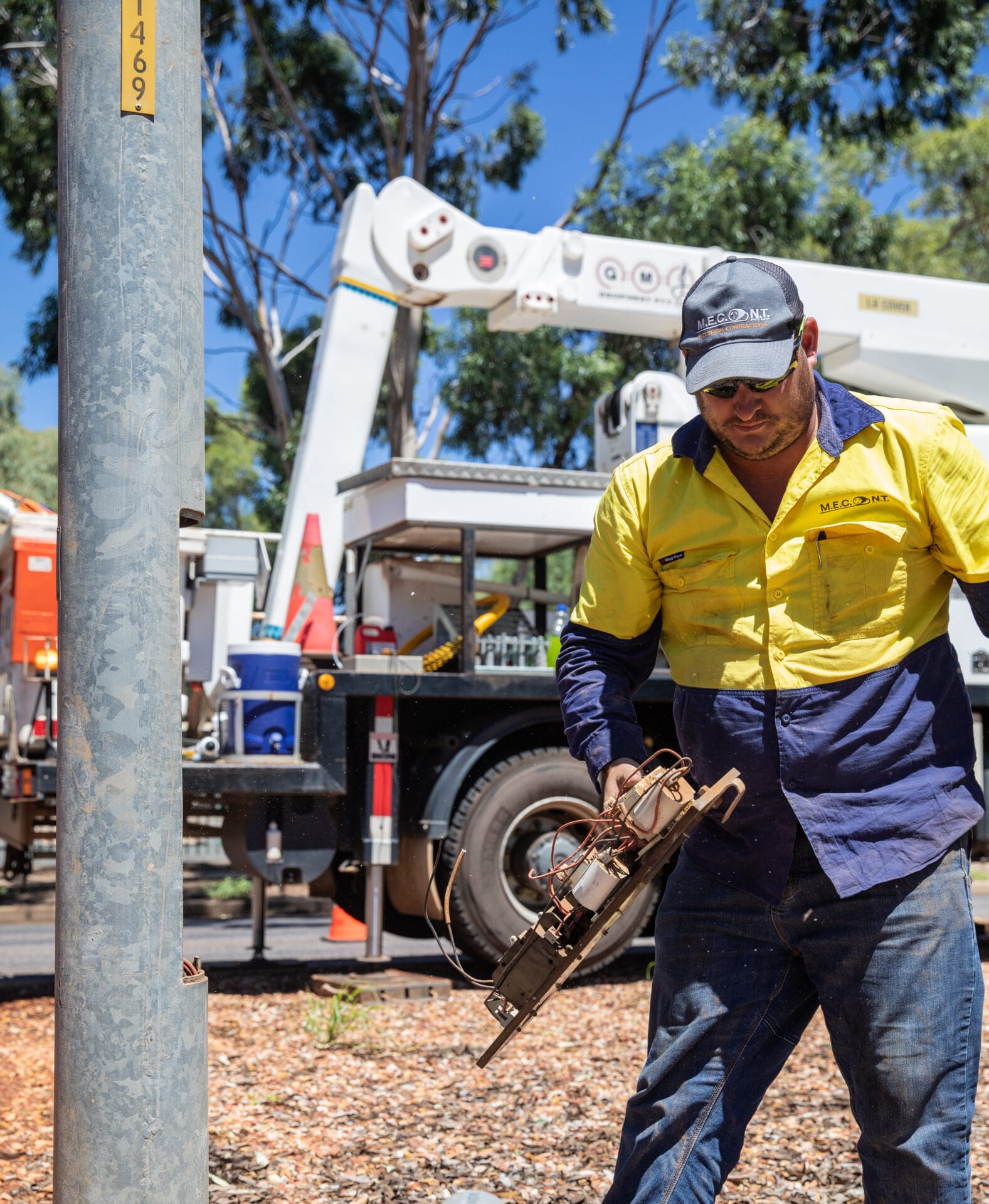 A worker in a yellow and navy uniform operates equipment near a utility pole, with a service truck and trees in the background.