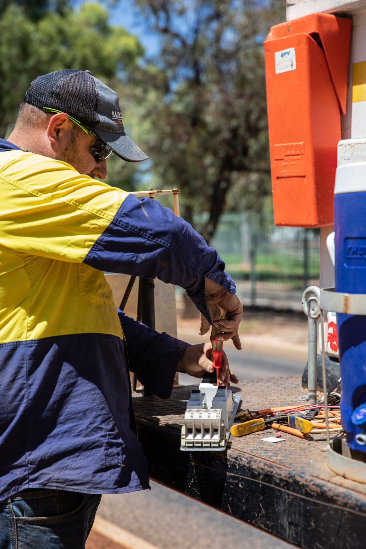 A worker in a high-visibility shirt uses a screwdriver to work on electrical equipment outdoors, with tools and supplies on the table nearby.