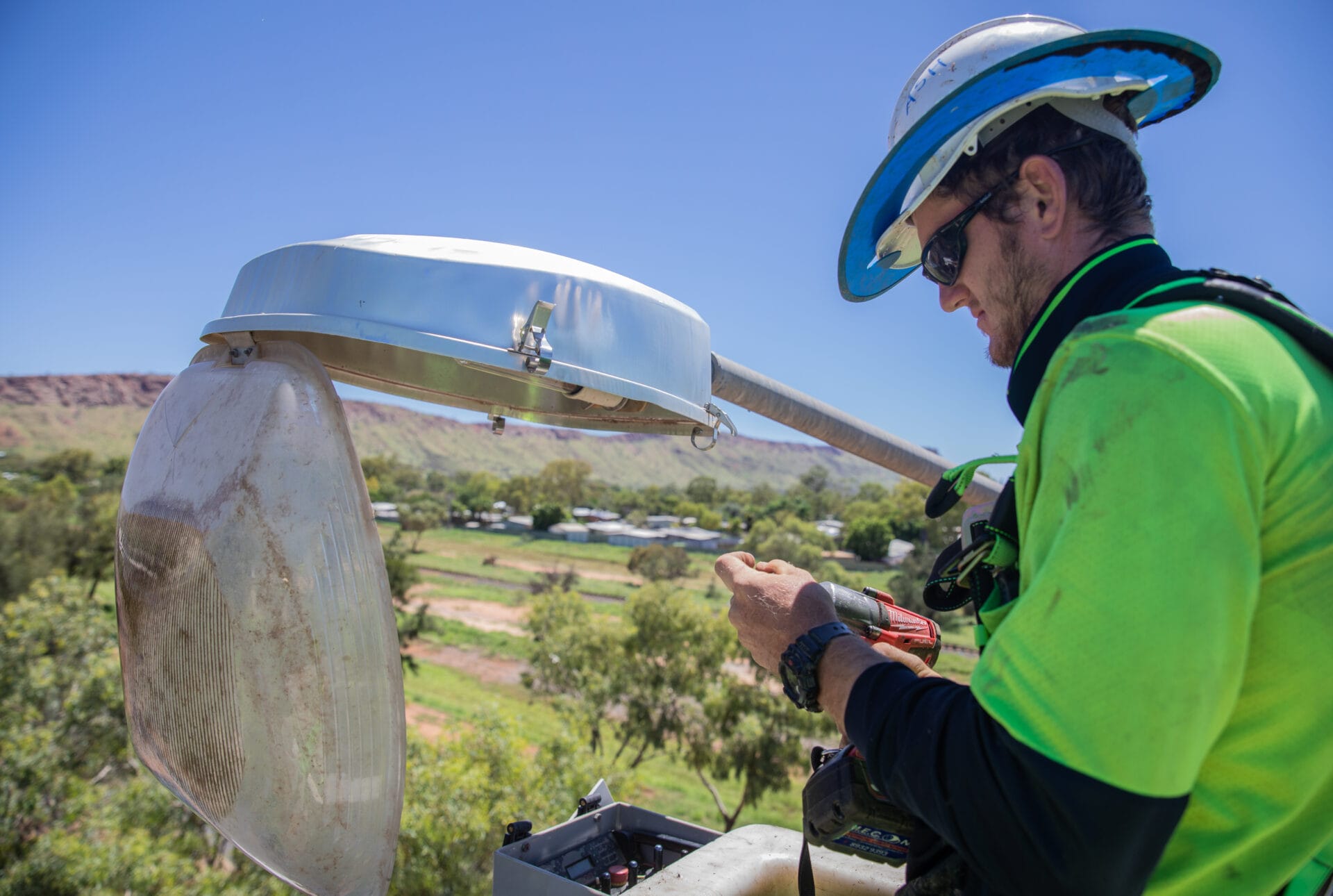 A utility worker in safety gear uses a tool to service equipment on a pole with a rural landscape in the background.