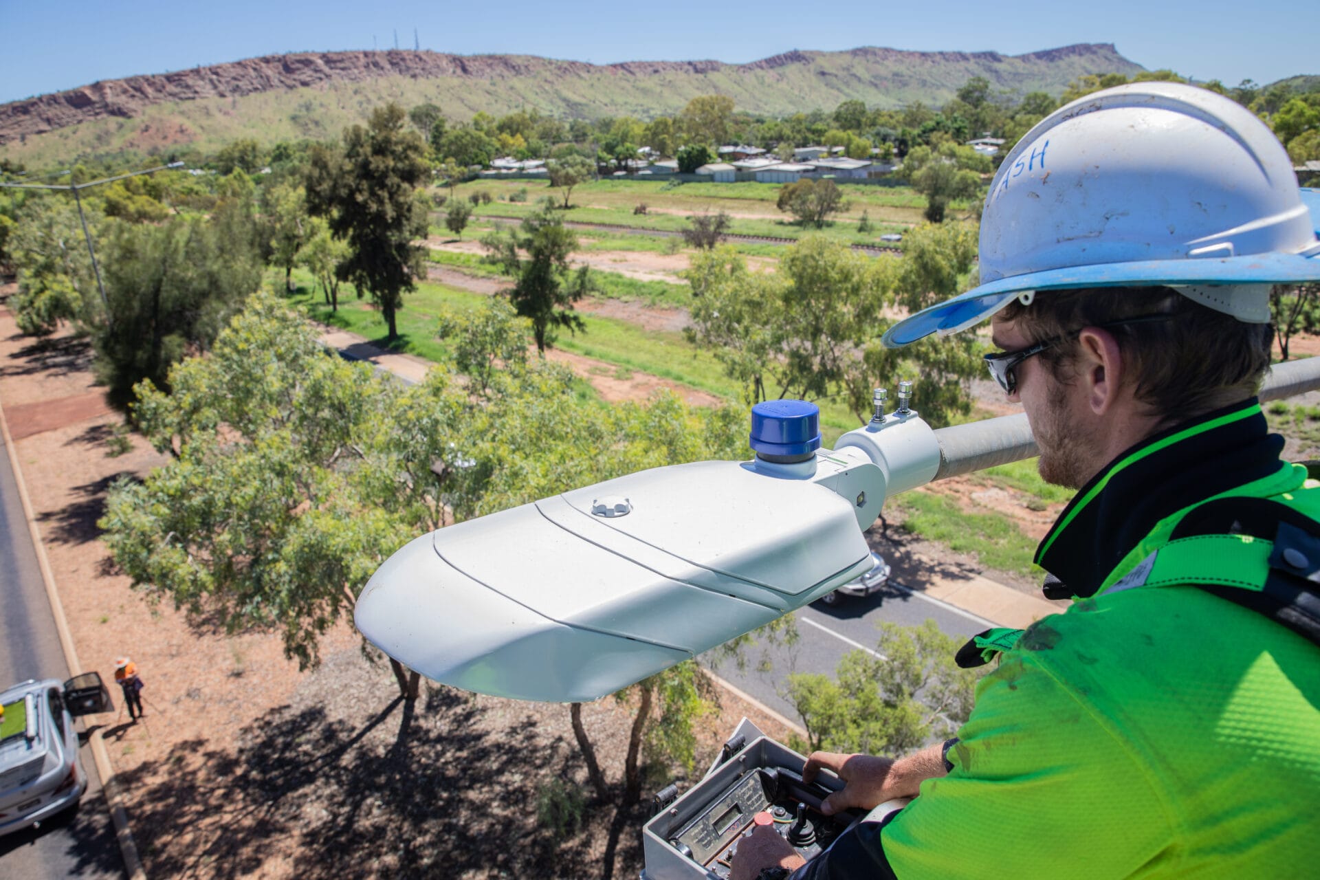 A worker in safety gear installs or maintains a streetlight from a raised platform overlooking a rural landscape with trees and distant hills.