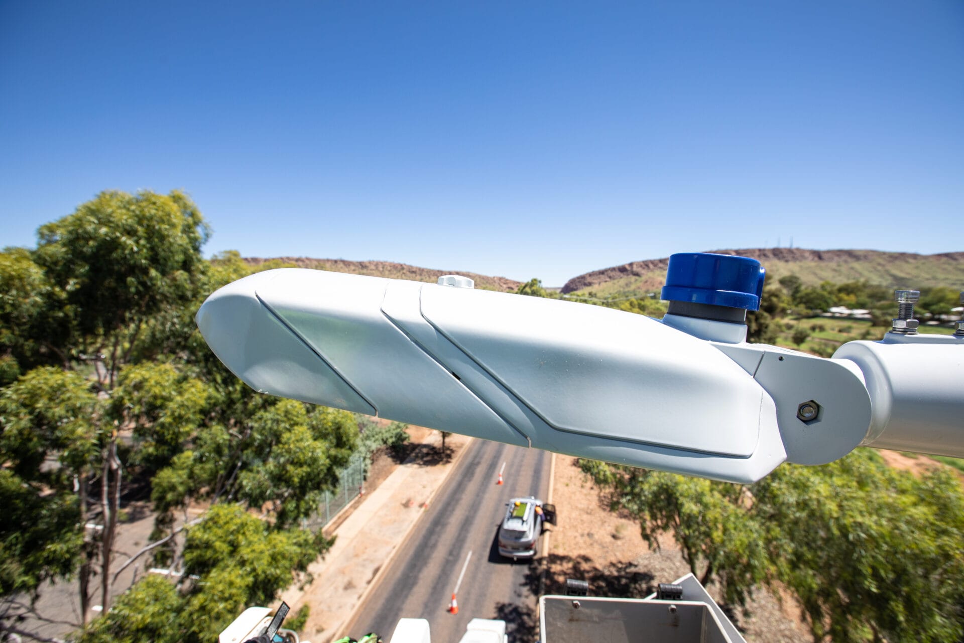 A close-up of a modern streetlight with a sensor mounted on a pole, overlooking a road lined with trees on a clear, sunny day.