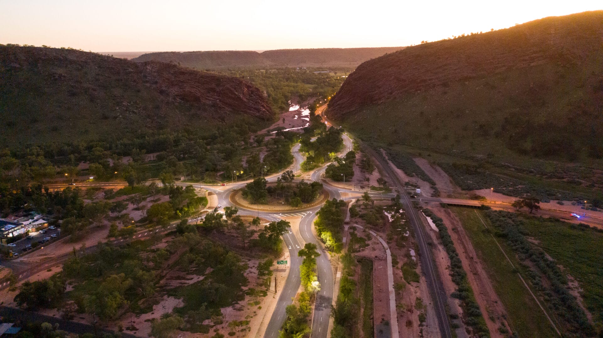 A winding road passes through a green valley between rocky hills at sunset, with lights illuminating parts of the road and a river visible in the distance.