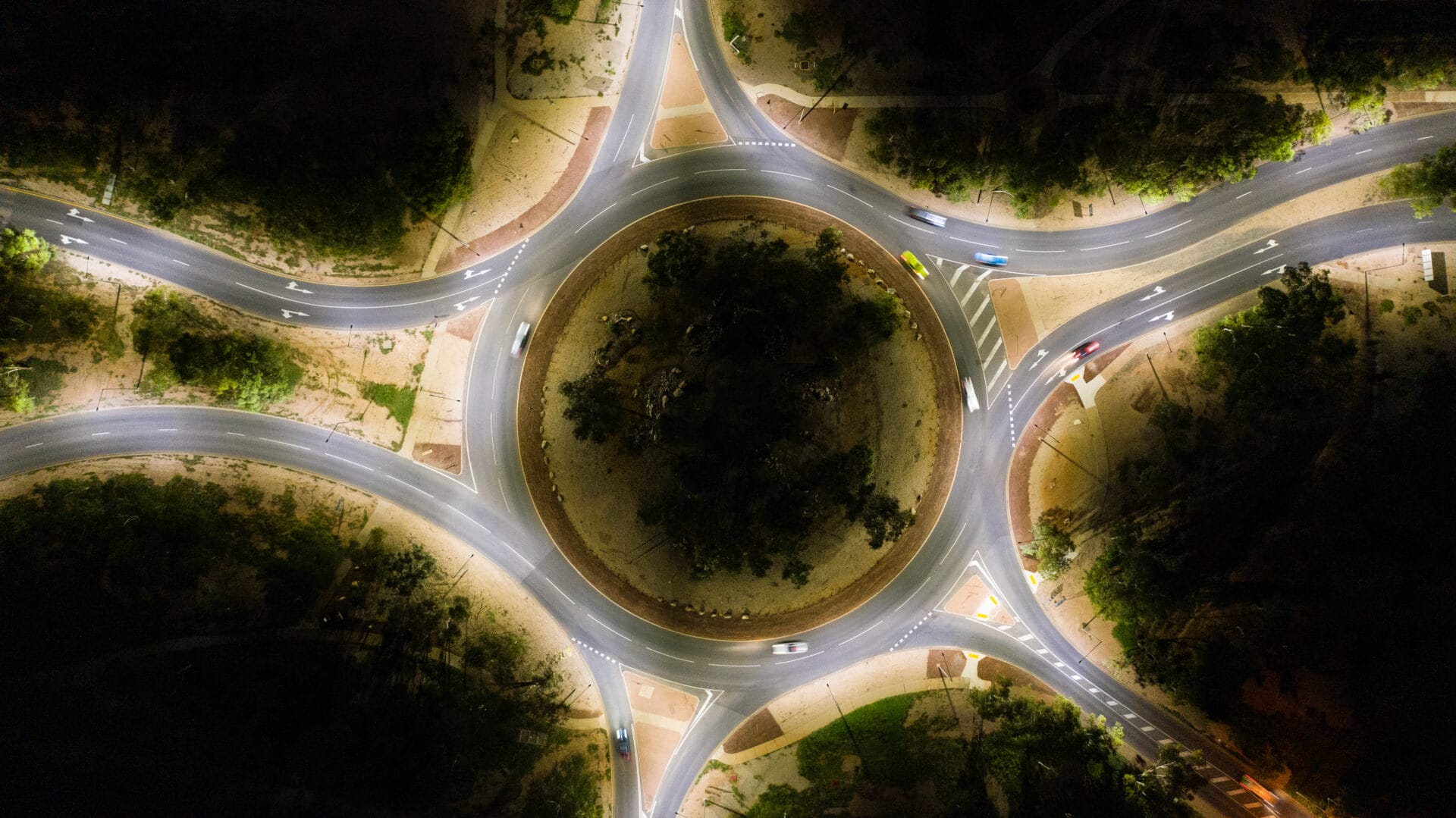Aerial night view of a circular roundabout with multiple roads branching out, surrounded by trees and illuminated by streetlights.
