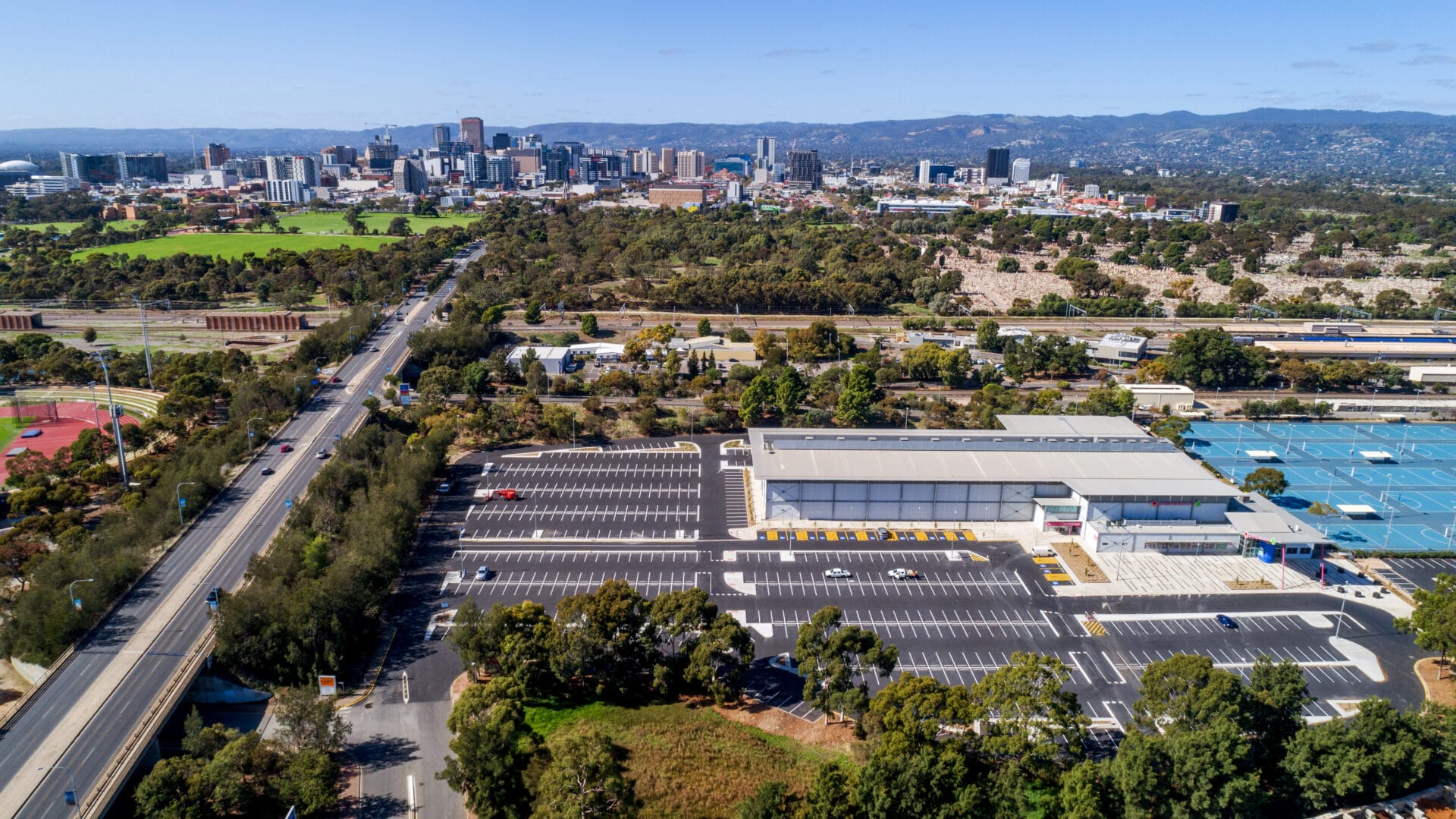 Aerial view of a large parking lot next to a sports facility, with a city skyline and green hills visible in the background.