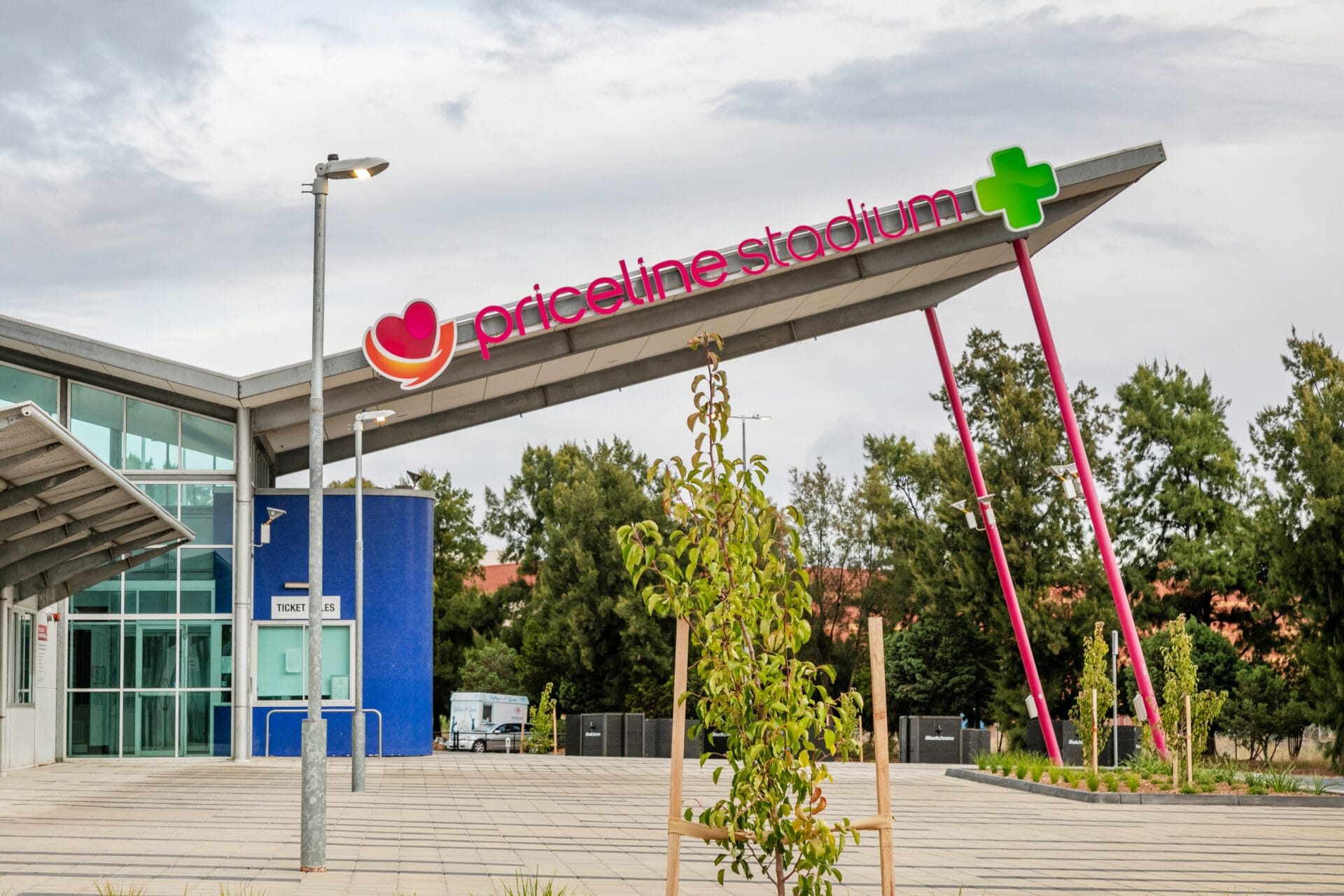 Entry of Priceline Stadium with modern angled roof, large pink and green signage, ticket office, and surrounding trees visible.