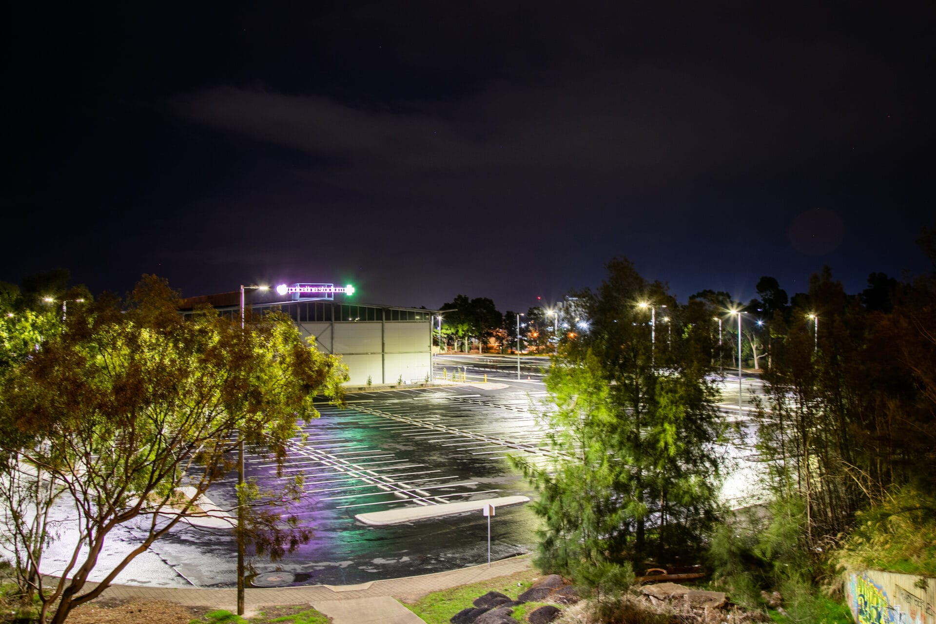 Empty, well-lit parking lot at night with a building in the background and trees framing the scene.