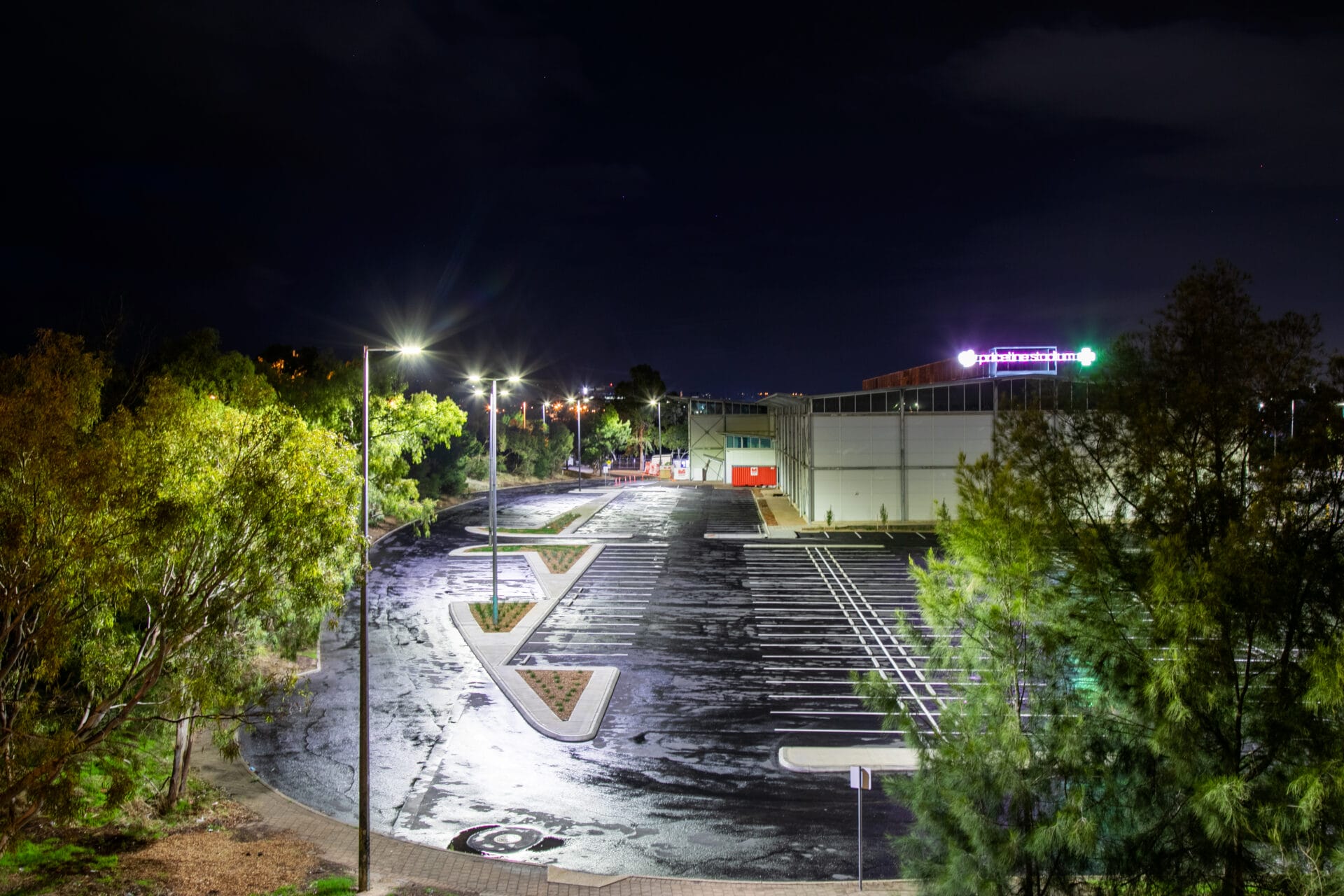 A mostly empty parking lot outside a large building at night, illuminated by streetlights, with trees surrounding the area.
