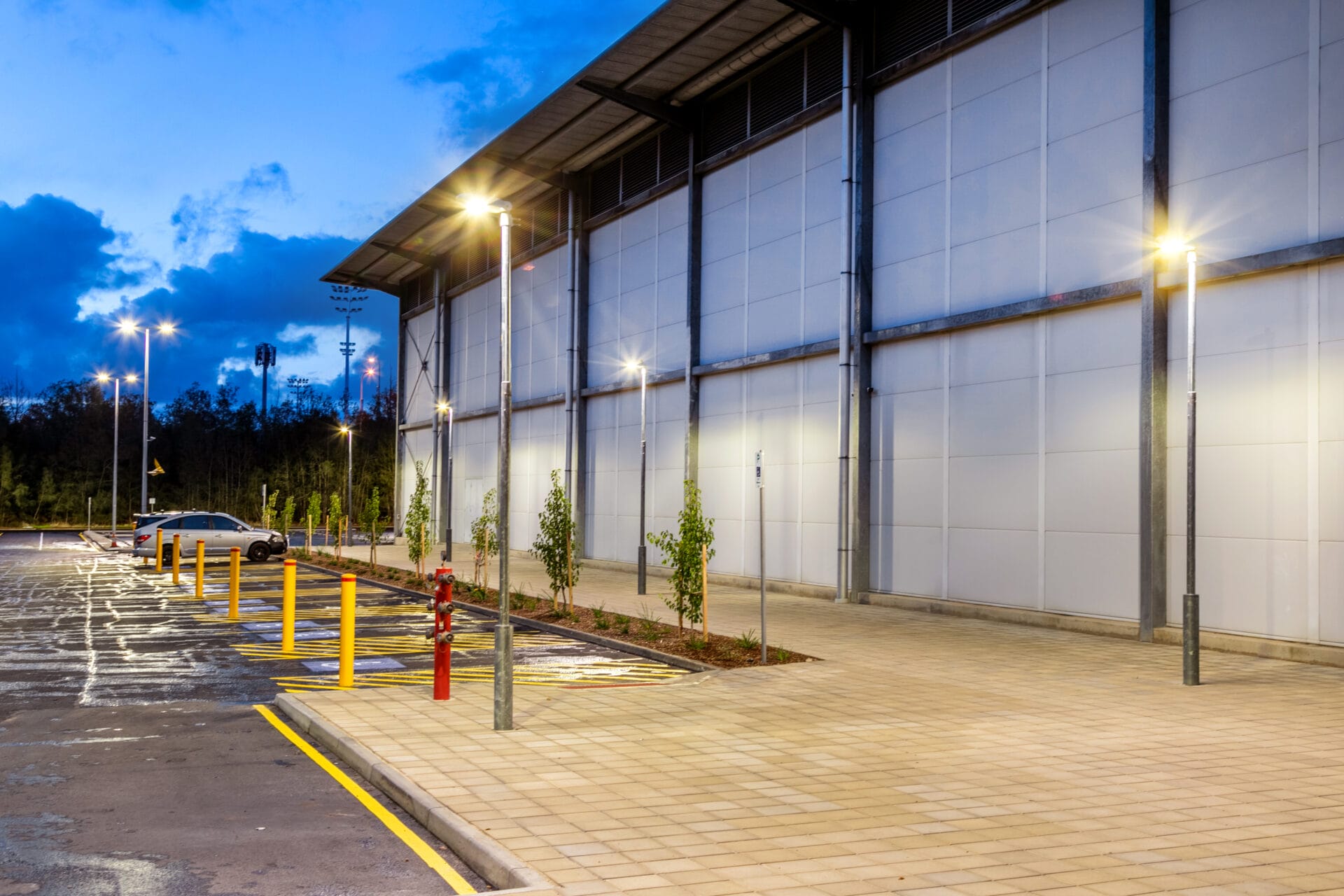 Empty parking lot with yellow bollards, sparse young trees, a single parked car, and a large industrial building at dusk with lights on.
