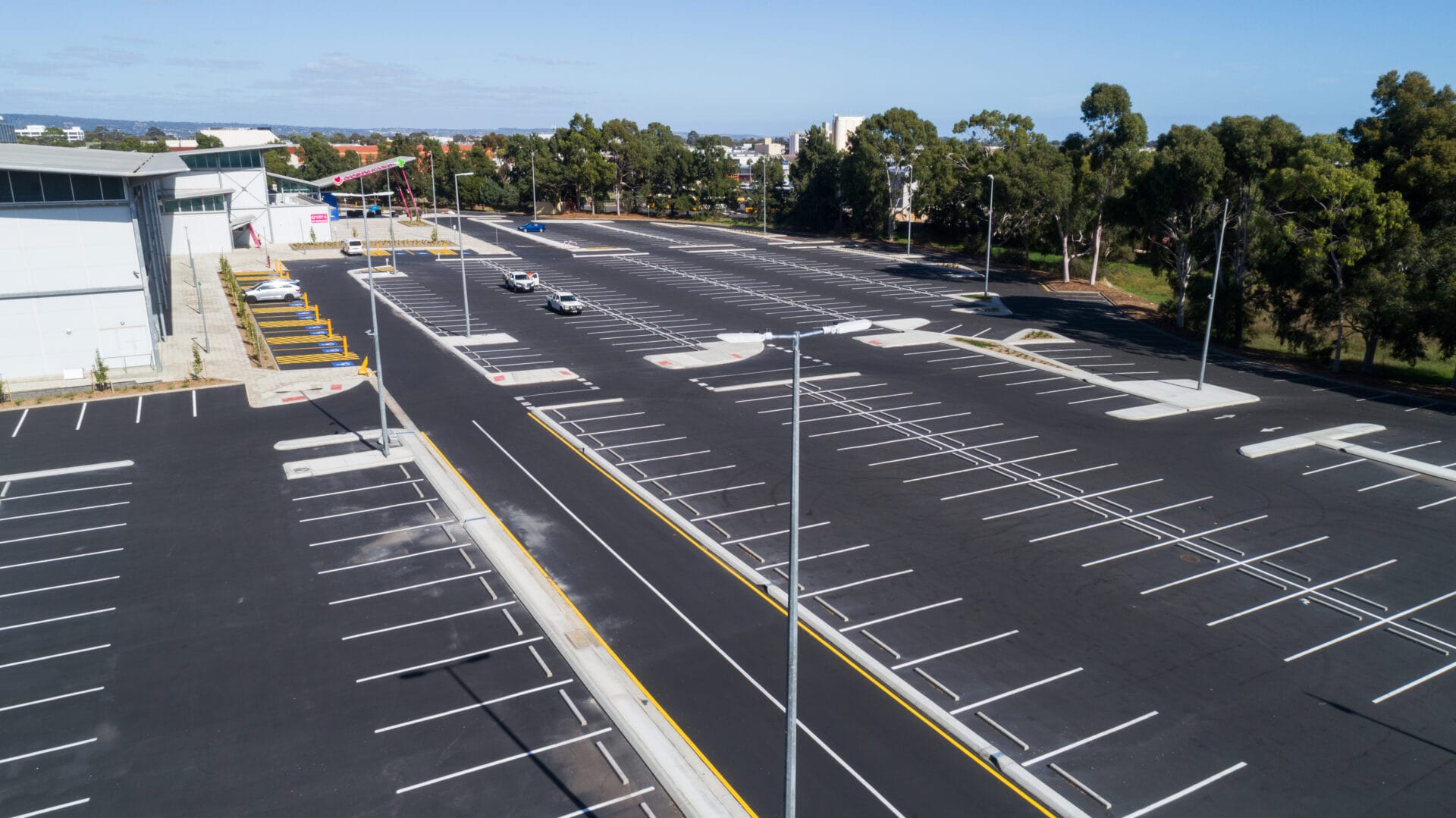 A large, mostly empty parking lot with a few cars parked near the center; surrounding trees and buildings are visible in the background.