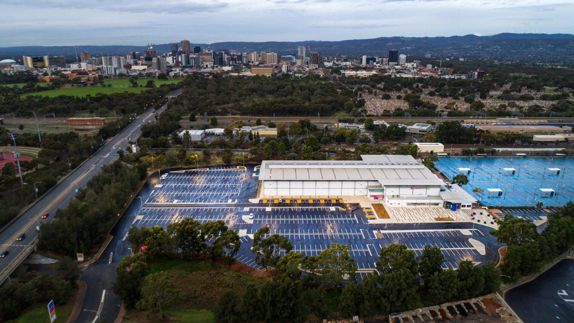 Aerial view of a large parking lot and commercial building surrounded by trees, with a city skyline and hills visible in the background under a cloudy sky.