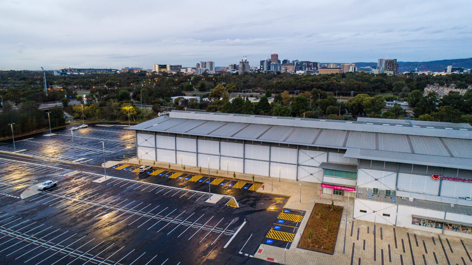 A large, mostly empty parking lot in front of a commercial building with a city skyline in the background on a cloudy day.