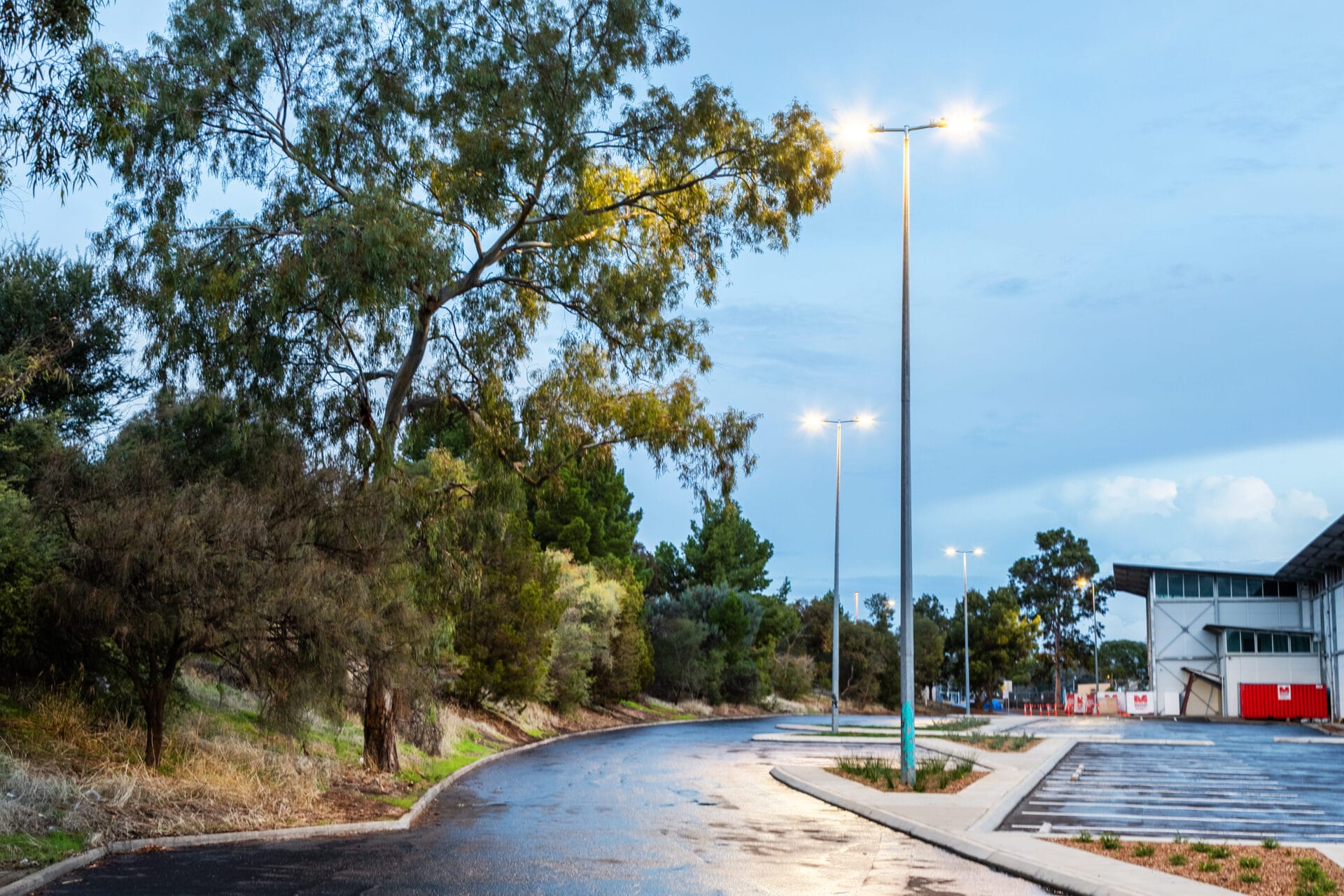 An empty parking lot next to a winding road lined with streetlights and trees, with a building visible on the right and a wet pavement reflecting light.